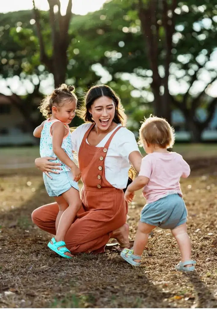 A beautiful young woman of Pacific Islander descent kneels down and lovingly interacts with her happy three year old daughter and one year old son while at the park in Hawaii.