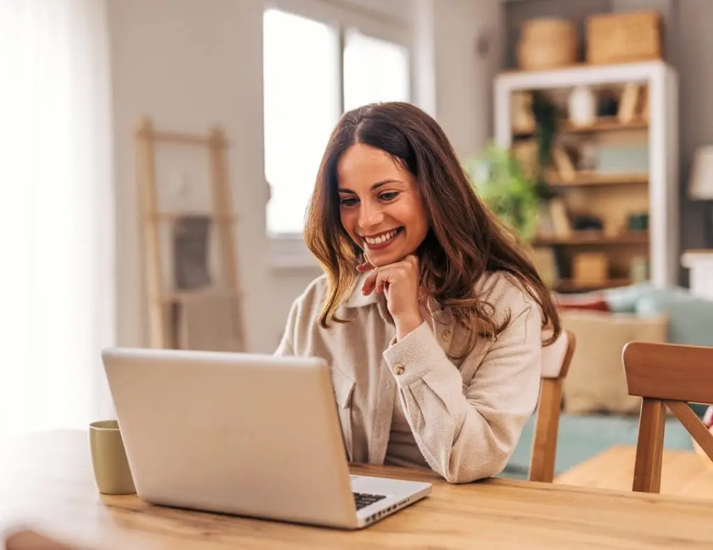 Happy young woman working from home, sitting at a dining table and smiling while looking at her laptop, enjoying the comfort of her bright living room filled with natural light